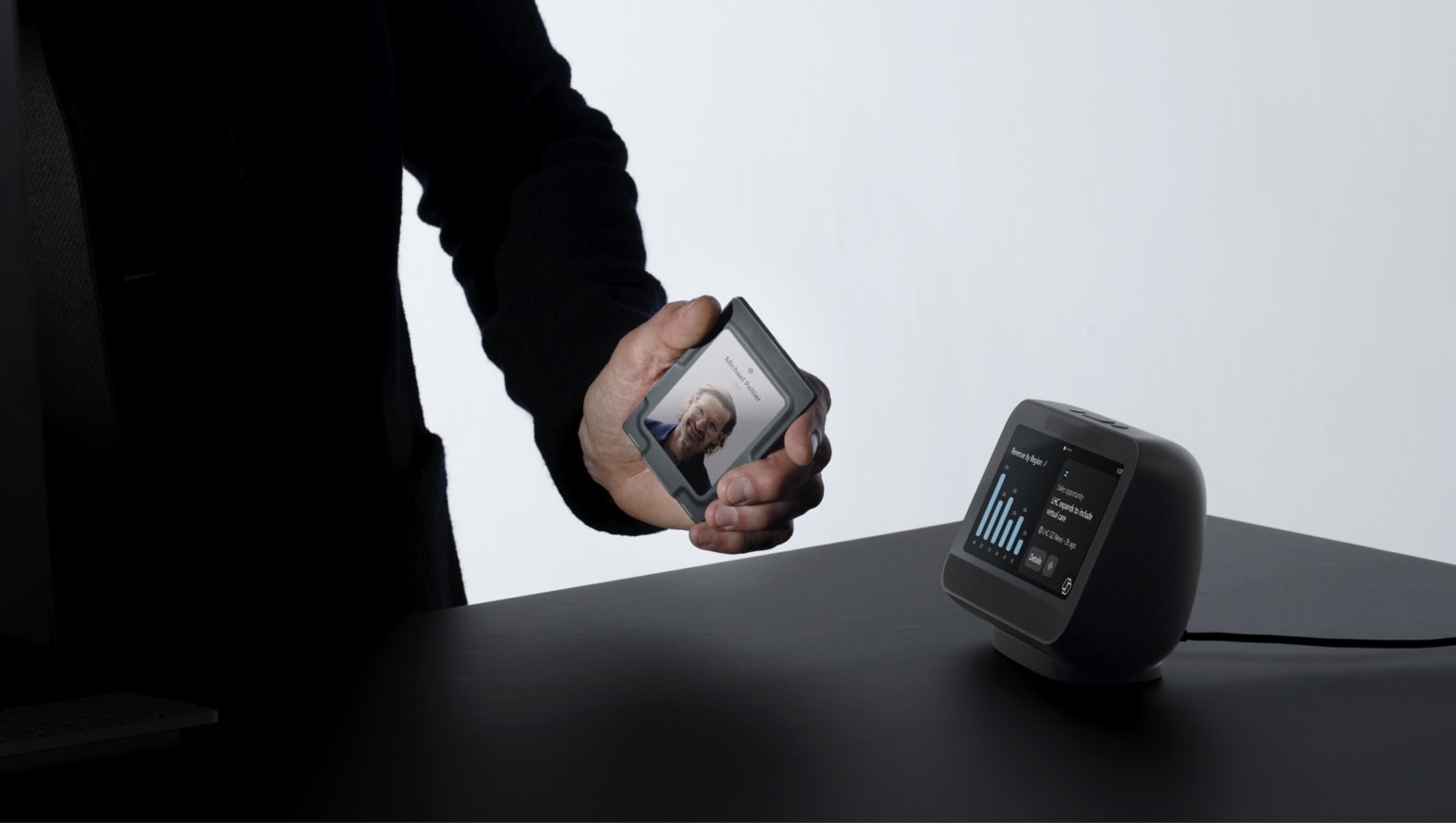 A badge held up beside the desk companion, the two devices showing the same work — handing a task between form factors